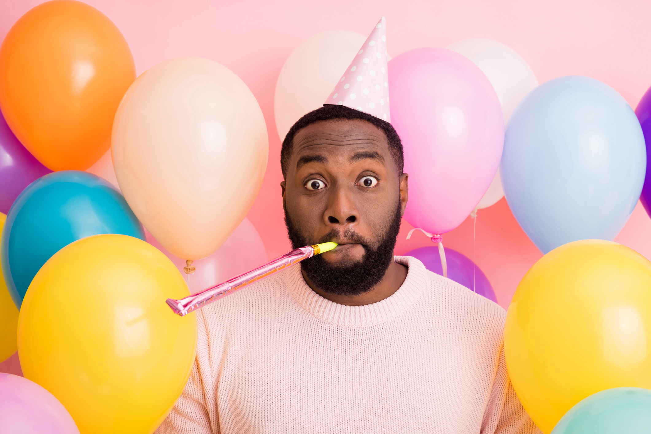 Photo of funky dark skin guy birthday surprise party surrounded many balloons, blowing noisemaker crazy guest wear paper cone cap sweater on pink color background